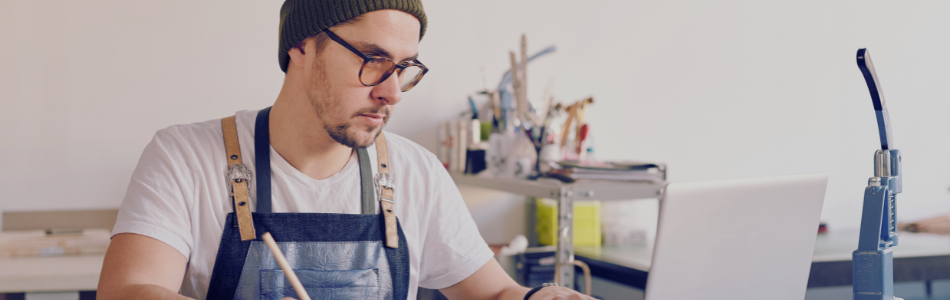 man in overalls looking at a laptop with an artistic workshop in the background
