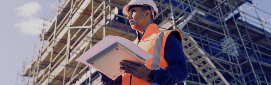Construction worker wearing a safety vest and holding a clipboard, building under construction in the background.
